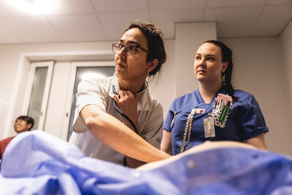 Två kvinnor som arbetar inom vården tittar åt vänster.  Den ena håller i ett stetoskop och har mörkt hår och glasögon. Den andra kvinnan har mörkt hår i en fläta.