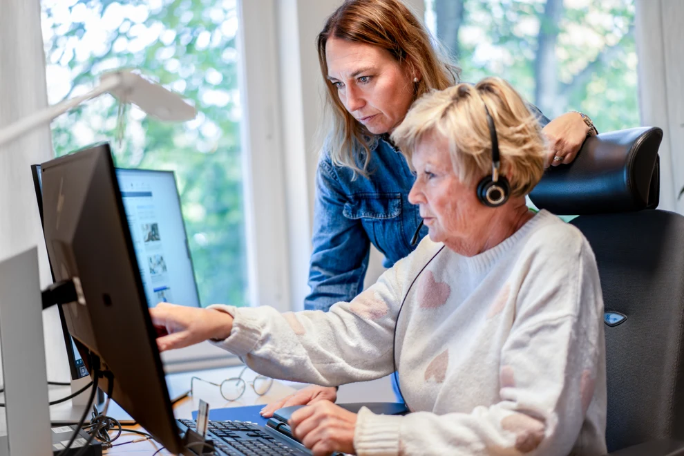 En kvinna med hörlurar sitter framför två dataskärmar. Hon pekar mot skärmen och visar något för en kvinna som står bredvid och tittar på skärmen.